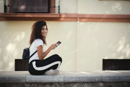 Young black woman with curly long hair sitting on top of the wall in urban place. Mixed girl wearing casual clothes and smiling while using her tablet device.の写真素材
