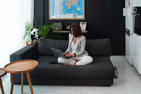 A middle-aged businesswoman sitting on a sofa with of her laptop and managing her online business remotely from home.の写真素材