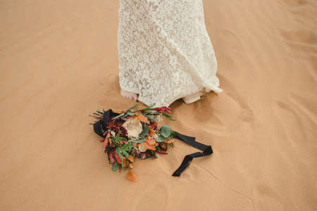 Beautiful bridal bouquet lying on the ground next to the bride's feet. Elegant flower bouquet outdoors on sand dune in desert. の写真素材