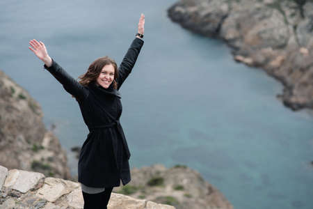 woman raises her arms to the sky as a sign of freedom on top of a cliff in front of the seaの写真素材