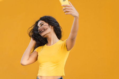 young mixed race woman inyellow t-shirt making a funny selfie with her phone with orange wall in the...