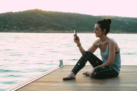 smiling young girl sitting on a pier making a video call with the headphonesの写真素材