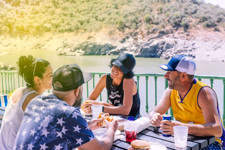 Group of four friends with hats to protect themselves from the sun on a sunny day sitting around a wooden table eating, drinking and having fun in the field with a river in the backgroundの写真素材