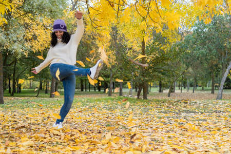 young woman in a hat and white wool sweater has fun kicking a pile of dry leaves that have fallen from the trees in a park in autumnの写真素材