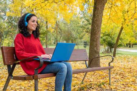 young woman with curly hair sitting on a bench working with laptop and blue wireless headphones while smiling in autumn with leaves falling from the treesの写真素材