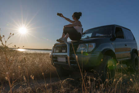 young woman taking a selfie with her mobile sitting on the hood of her off-road car while on a lake in summer on vacationの写真素材