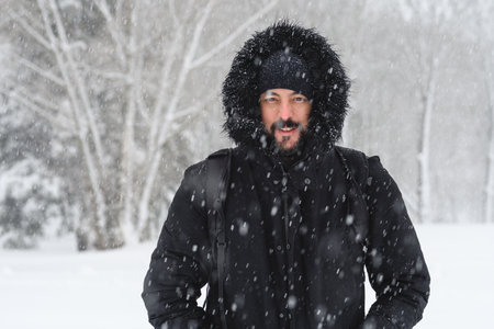 portrait of a young hipster man with a beard wearing a coat and hat from the cold while it is snowing in winter in a park with snowy trees in the backgroundの写真素材