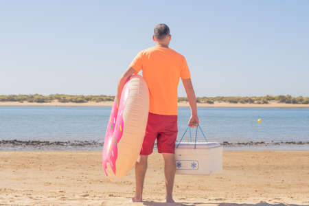 Young hipster man with a beard, swimming trunks and sunglasses carries a pink inflatable float and a fridge on the beach. leisure on a sunny day on vacationの写真素材