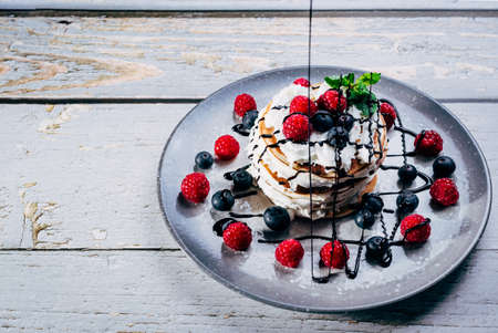 colorful plate of pancakes with cream, mint leaves and fresh berries with chocolate syrup falling on top on wooden tableの写真素材