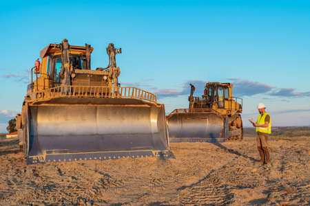 Construction inspector wearing safety helmet and reflective vest taking notes during an inspection of heavy machinery on a road construction siteの写真素材