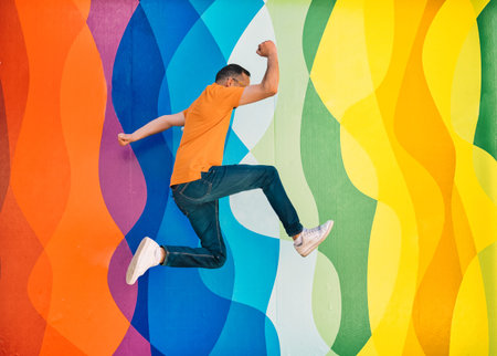 young man in orange polo shirt, jeans and sunglasses has fun while jumping on a colorful wall....