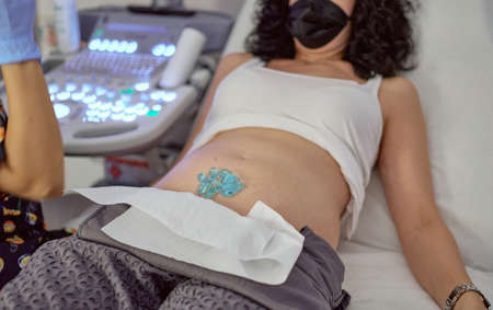 pregnant woman with face protection mask due to the pandemic,  with conductive liquid in her belly prepared to have a control ultrasound in a clinicの写真素材