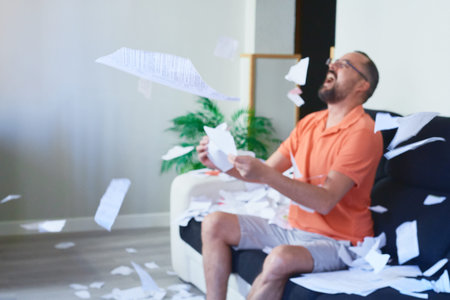 A young hipster man with a beard sitting on the sofa at home throwing papers in the air while...
