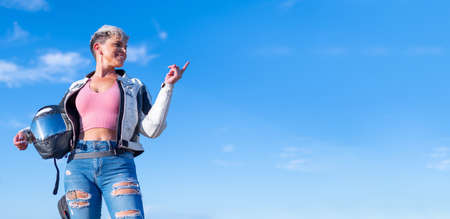 Smiling pretty young woman with short blond hair dressed as a biker with ripped jeans holding her motorcycle helmet points up with her finger. blue sky background. copy spaceの写真素材