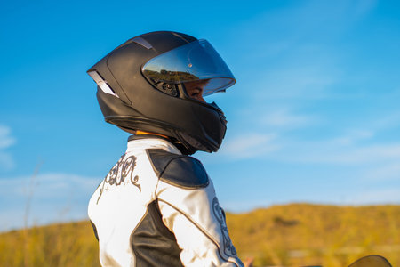young biker woman in profile dressed in white patterned jacket and black safety helmet. blue sky backgroundの写真素材