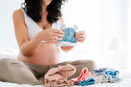 young pregnant woman with curly hair sitting on the bed looking at a little hat for her future...