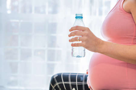 pregnant young woman in pink t-shirt holding in her hand a bottle of water to hydrate after exercising. concept health care and hydration in pregnancyの写真素材