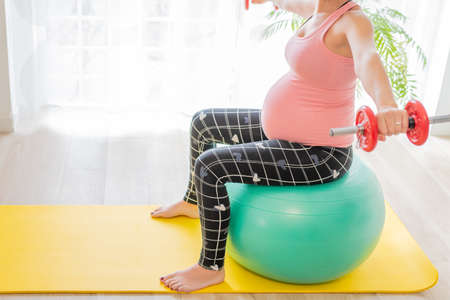 young pregnant woman lifting dumbbells sitting on a fitness ball. body toning and exercises during pregnancyの写真素材