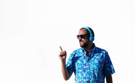 young hipster man with beard, sunglasses, hawaiian shirt and blue headphones smiling points finger up. copyspace. white backgroundの写真素材