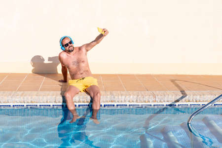 smiling young hipster man with beard, yellow swimsuit and sunglasses takes a selfie with his smartphone in the pool with his feet in the water while listening to music with his blue headphones.の写真素材
