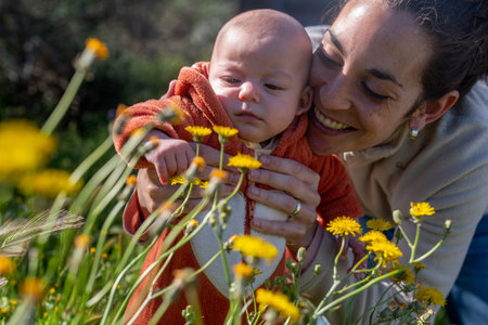 young smiling mother with her baby in the field touching the yellow flowers while going for a walk...