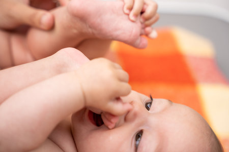 little smiling baby lying on a blanket putting her feet in her mouth due to discomfort caused by teething. concept of the emergence of the first teeth in the childhood stageの写真素材