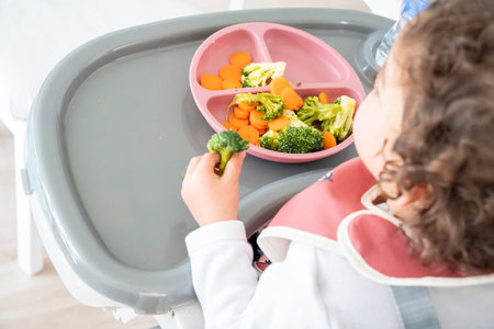 Back view image of a curly-haired baby girl sitting in her highchair eating a plate of vegetables. Baby-led weaning concept, healthy and balanced diet. Healthy lifestyle habits.の写真素材