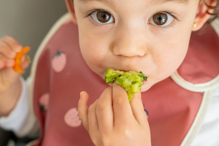 little girl sitting in her high chair enjoying eating vegetables, holding broccoli in her hand....