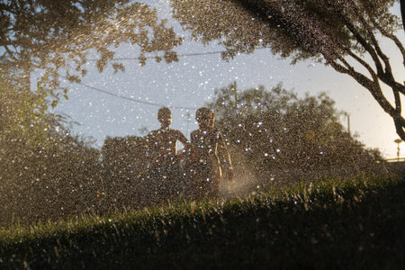 Silhouettes of children who play with the water that comes out of the sprinklers to water a garden...