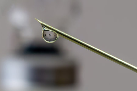 Drop of vaccine against Covid-19 (SARS-COV2) coming out of a syringe. A vial of vaccine is reflected...