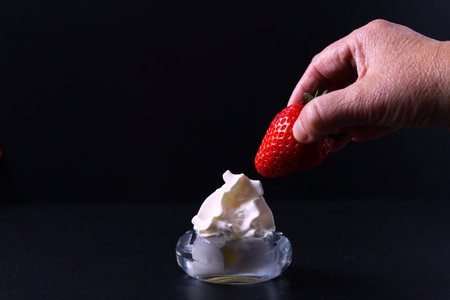 A woman's hand is about to spread a strawberry with whipped cream that is in a clear glass bowl. All with a black background.の写真素材