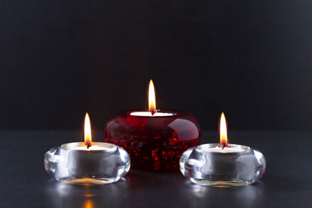 Mourning or prayer concept. Three burning candles on black background. Two of them in transparent glass candlesticks and one in red glass chandelier.の写真素材