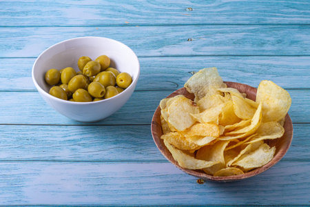 Spanish snack concept. Stuffed olives in a white bowl and potato chips in a wooden bowl on a wooden table in blue tonesの写真素材