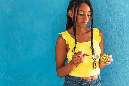 black afro latin american healthy girl with braids eating a kiwi with a spoon. Yellow shirt and blue jeans skirt. Blue wall in the background, turquoise. Beautiful skinny woman ,green yellow make upの写真素材