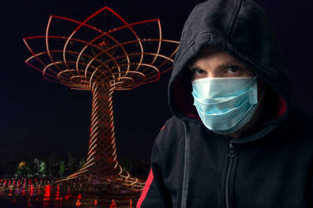 Portrait of a cloaked man wearing a surgical respiratory mask against coronavirus COVID-19 viral infection. Background is a landmark of Milan at night.の写真素材