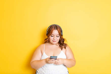 Hispanic fat young woman using smart phone and headphones on isolated yellow background. mexicanの写真素材
