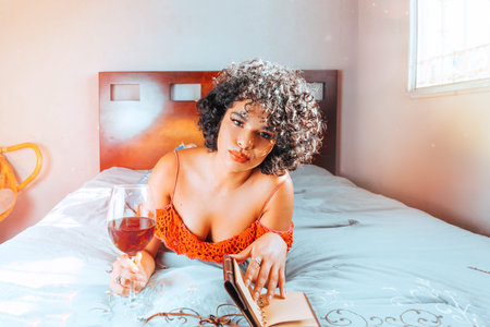 beautiful brunette hispanic woman lying in bed reading book with glass of rose wine