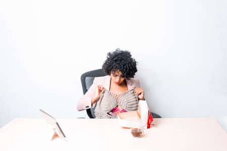 young hispanic woman taking clothes out of gift box on video call in front of laptopの写真素材
