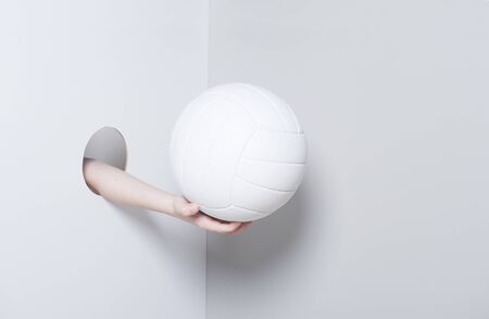 Hand in the hole. A kid hand holds a white volleyball ball on grey and white backgroundの写真素材