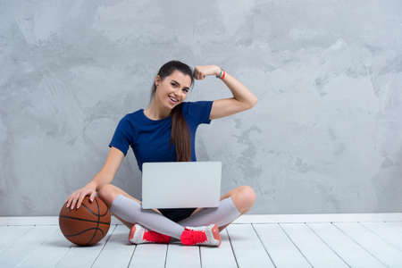 Young girl doing exercises with basketball ball watching online tutorials on laptop. A sports coach leads an online lesson. Online workout concept.の写真素材