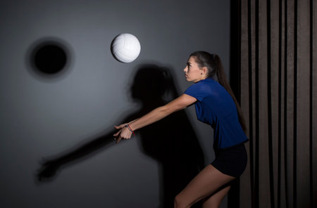 Volleyball girl hold and kick ball in blue costume on dark wall background. Player doing sport workout at home. Sport and recreation conceptの写真素材