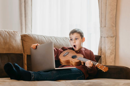Boy playing ukulele and watching online course on laptop while practicing at home. Online training, online classes.Vintage color filterの写真素材