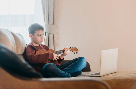 Boy playing ukulele and watching online course on laptop while practicing at home. Online training, online classes.Vintage color filterの写真素材