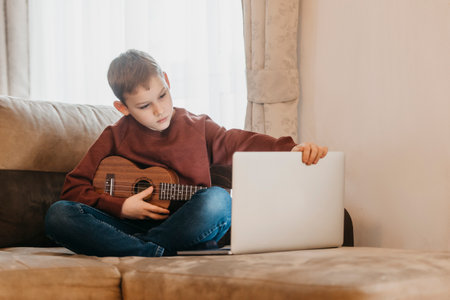 Boy playing ukulele and watching online course on laptop while practicing at home. Online training, online classes.Vintage color filterの写真素材