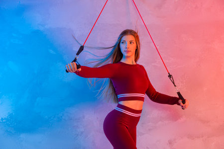 Female fitness instructor working out with a rubber resistance band in blue and pink neon light in gym. Sport online conceptの写真素材