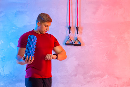 Athletic trainer with blue massager and rubber resistance on a bright neon background. Fitness man looking at her wrist watch. Interactive coach conceptの写真素材