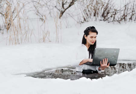Girl with laptop in frozen lake ice hole. Woman hardening the body in cold water.の写真素材