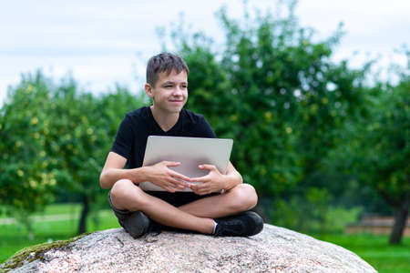 Boy sitting on a stone with laptop in garden. Kid have online web lesson or class on computer at nature, green class. Back to school. Outdoor learning conceptの写真素材