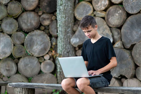Boy sitting on wooden seat with laptop in forest. Kid have online web lesson or class on computer at nature, green class. Back to school. Outdoor learning conceptの写真素材
