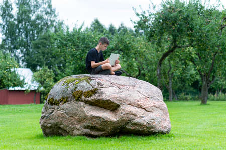 Boy sitting on a stone with laptop in garden. Kid have online web lesson or class on computer at nature, green class. Back to school. Outdoor learning conceptの写真素材